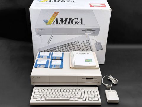 Amiga 1000 console with its keyboard and mouse in front, four blue floppy disks and the Amiga BASIC manual on top, and the original Amiga retail box standing behind it