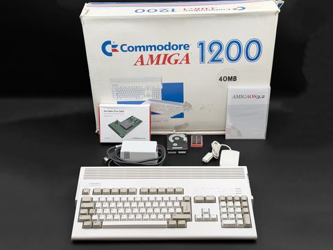 A Commodore A1200 case with keyboard sits in the foreground in front of its original 'Commodore Amiga 1200 40MB' retail box, with a Terrible Fire 1260 retail box, an AmigaOS 3.2 CD case, a 2.5-inch hard drive, a pair of chips on pink foam, the power supply and a mouse arranged between them