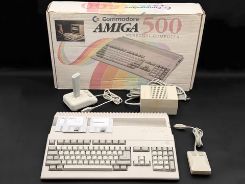 A Commodore Amiga 500 sits in front of its original 'Commodore Amiga 500 Personal Computer' box, with a white joystick, the power supply, a two-button mouse and three 3.5-inch floppies (one labeled 'Datastorm') laid out around it