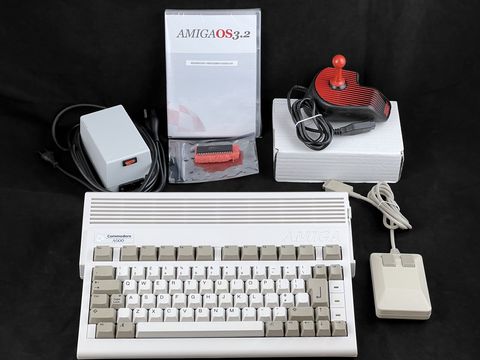 Amiga 600 with mouse and a red-handled joystick in front, a gray power supply at left, an AmigaOS 3.2 package and a white box behind