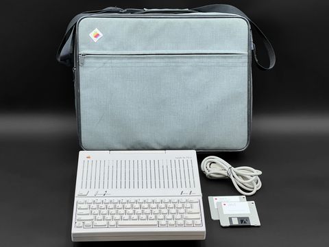A grey Apple IIc Plus sits at the front with its power cord and two 3.5-inch floppy disks beside it, with its grey fabric carrying bag standing behind shot straight on
