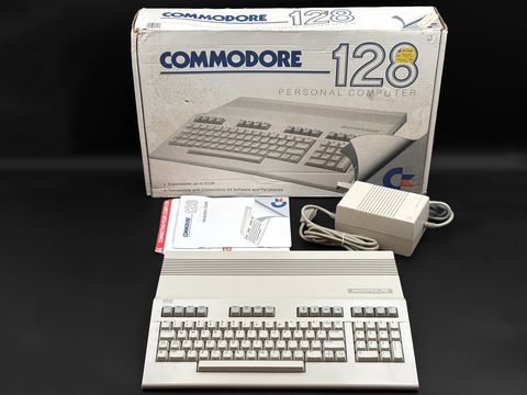 A Commodore 128 keyboard unit sits in front of its open-faced Commodore 128 retail box, with a manual and a beige power supply with cables