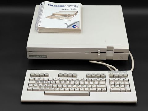 A Commodore 128D main unit with the Commodore 128D System Guide resting on top, its detachable keyboard laid in front connected by a cable