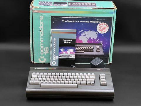Commodore 16 computer straight on in front of its original teal box, with the Owner's Guide and black power supply beside it