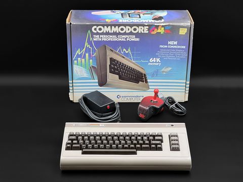 Commodore 64 breadbin in front of its original retail box with a power supply and a red joystick placed between them