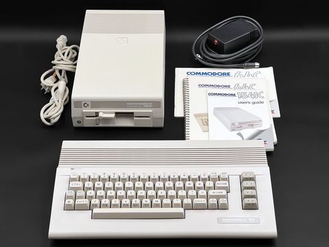 A Commodore 64C keyboard unit sits in front of a Commodore 1541C disk drive, with a black power supply, an off-white power cord, and the 64C and 1541C user's guides arranged, shot from above