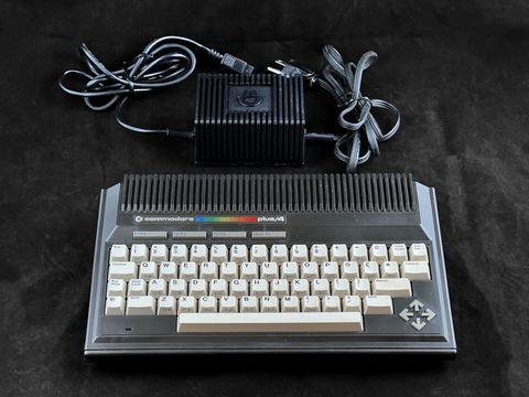 Commodore Plus/4 viewed from slightly above on the dark backdrop with its black power supply and cord coiled behind it
