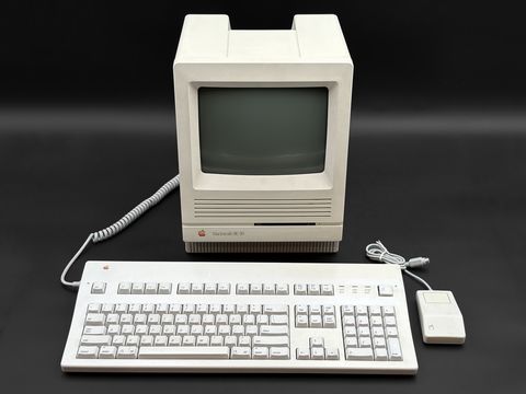 Front view of a Macintosh SE/30 (screen off) with an Apple Extended-style keyboard and mouse laid out in front of it, coiled keyboard cable at left