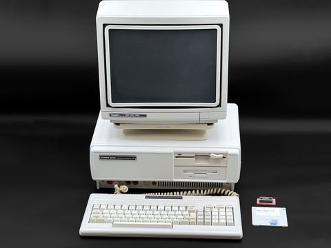 Tandy 1000TX desktop with a Tandy color monitor on top and the keyboard in front, with a small accessory and a manual or disk sleeve at the right, all straight on against a black backdrop