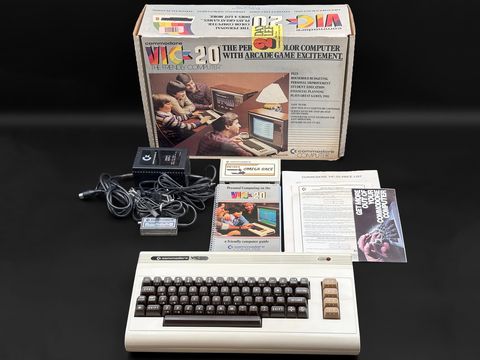 VIC-20 computer on the dark backdrop in front of its original illustrated retail box, with the power supply and cables, RF modulator, user guide and paperwork laid out between them
